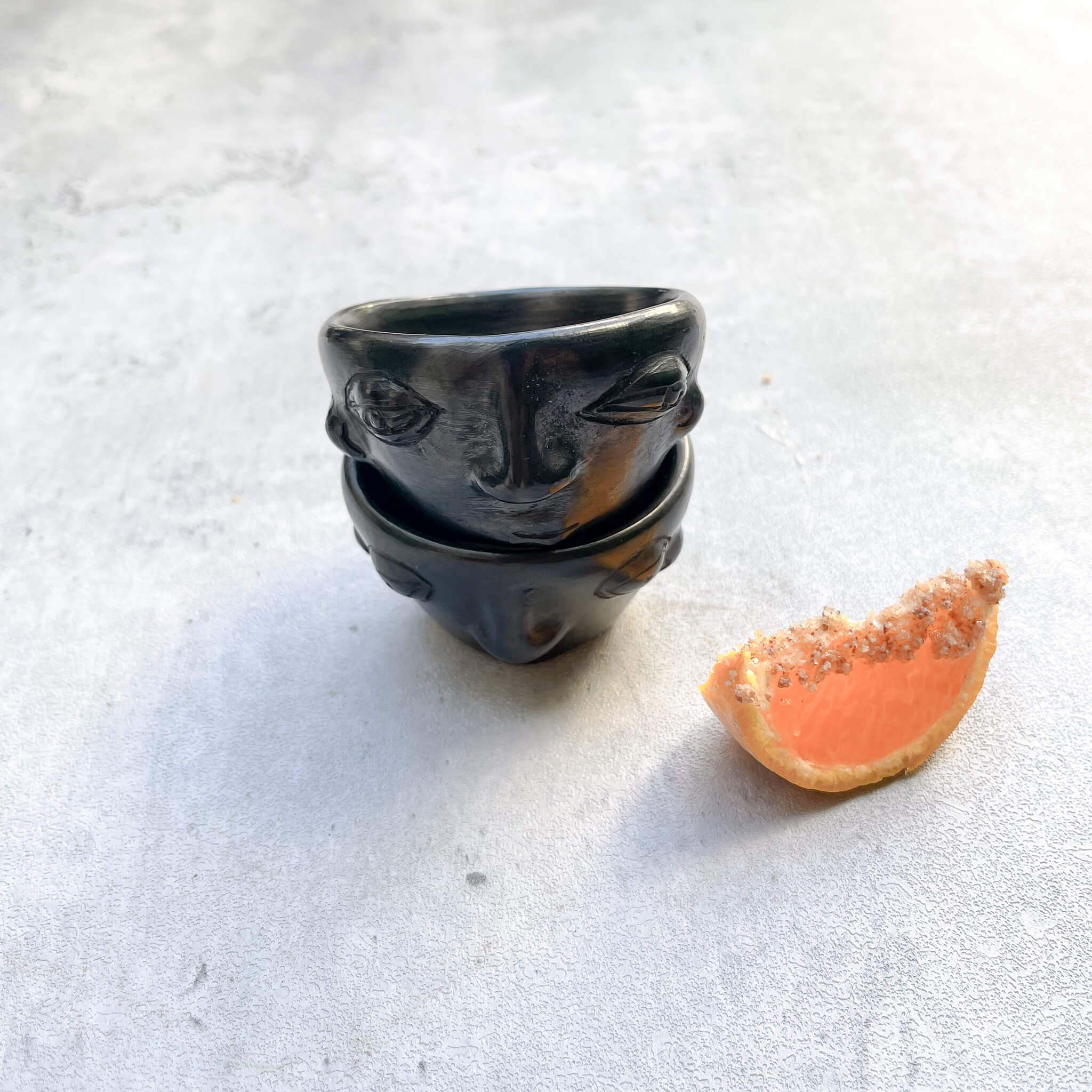 A pair of stacked black clay mezcal copitas next to an orange slice with salt.