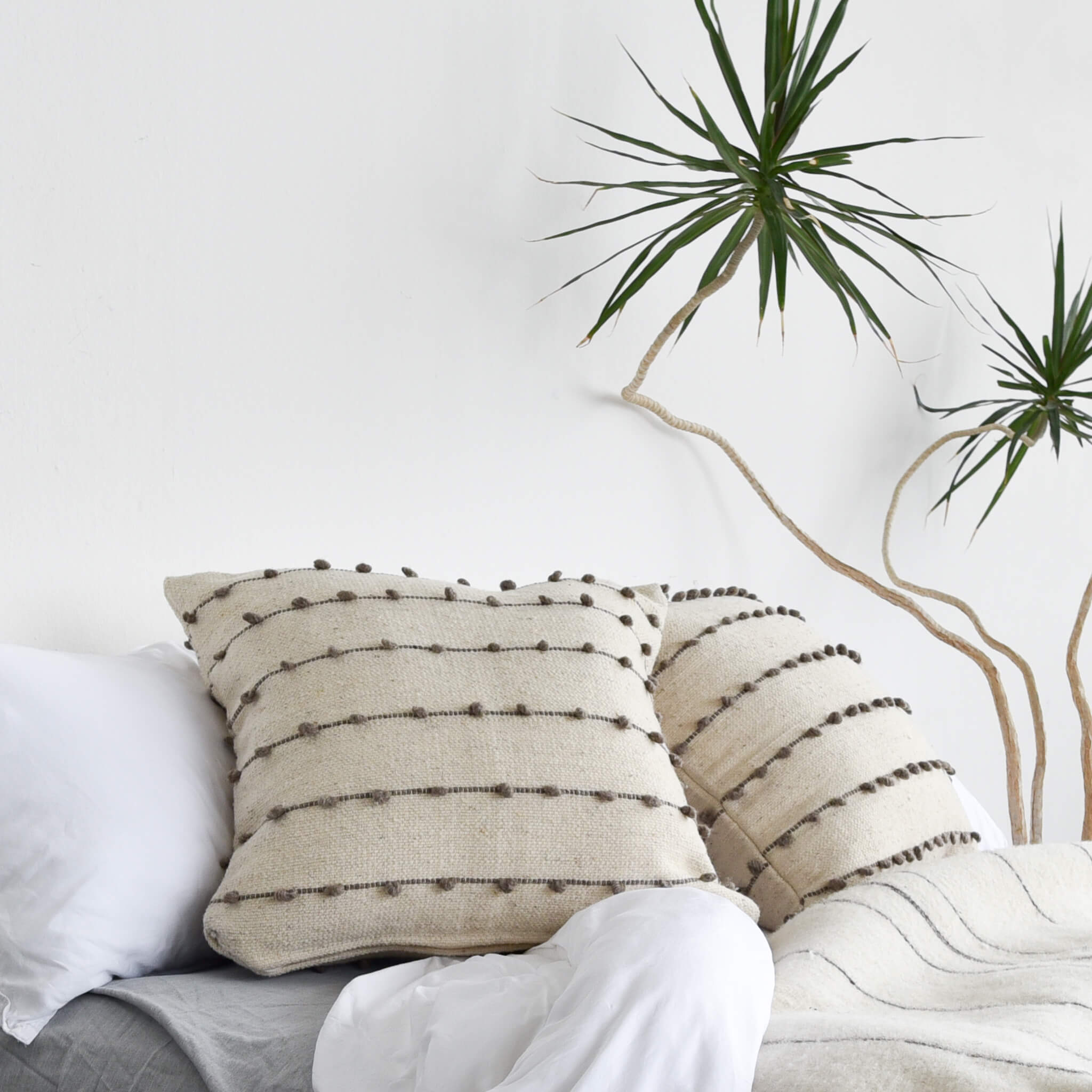 La Borla wool throw pillows with gray stitching on a white bed next to a sculptural yucca plant.