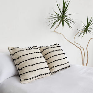 La Borla wool throw pillows with black stitching on a white bed next to a sculptural yucca plant.