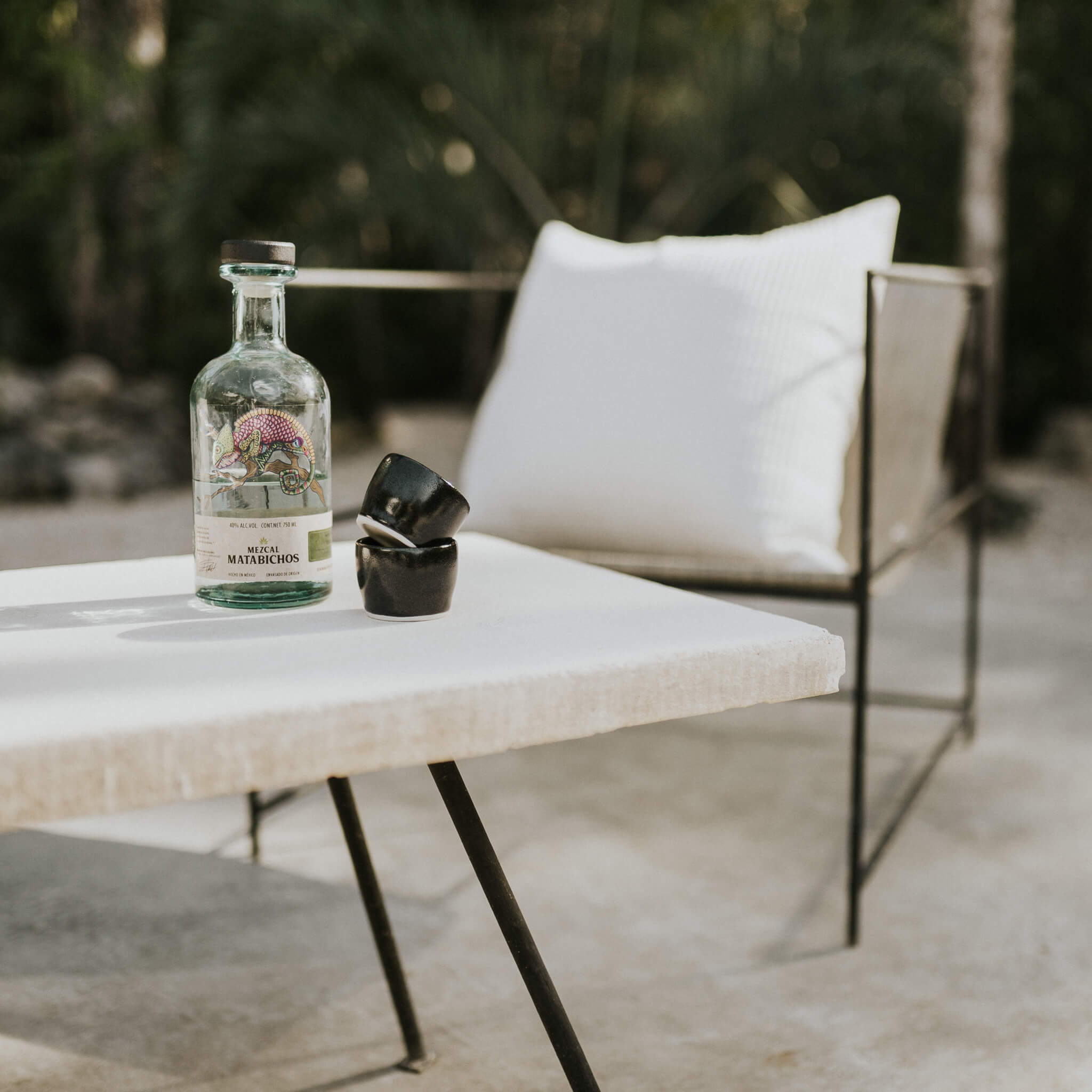 A set of black stoneware mezcal copitas on an outdoor table with a bottle of mezcal.