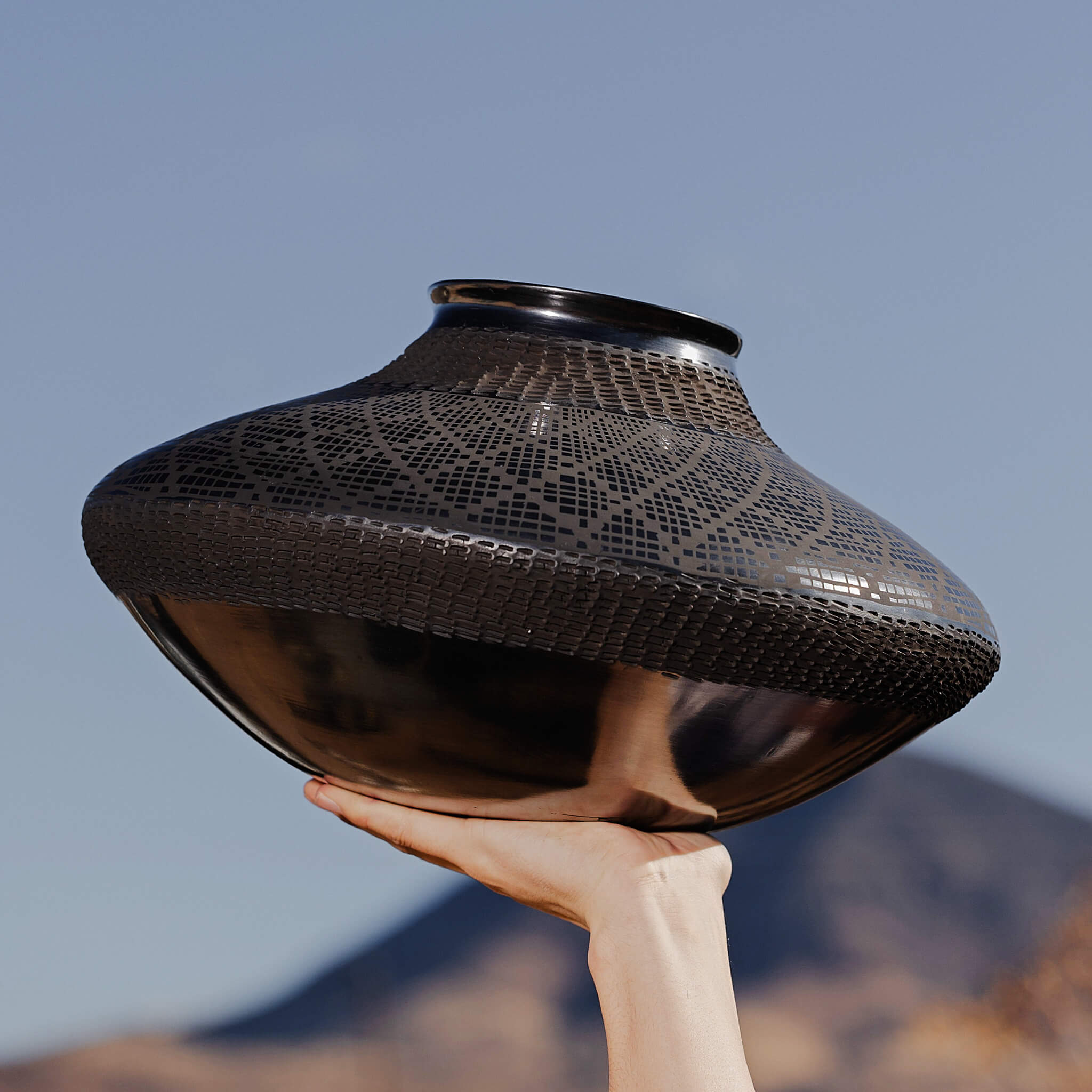 A large Mata Ortiz spheroid shaped vase held in a hand against a mountainous backdrop.