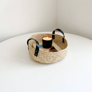 A medium Oaxaca palm storage basket with black leather handles holding a burning candle atop a book.
