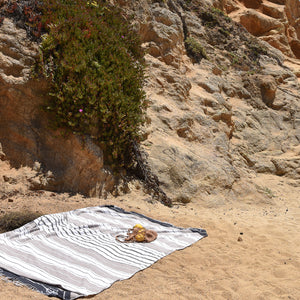 A cotton blanket handwoven in Mexico on a sandy beach with dramatic stone cliffs.