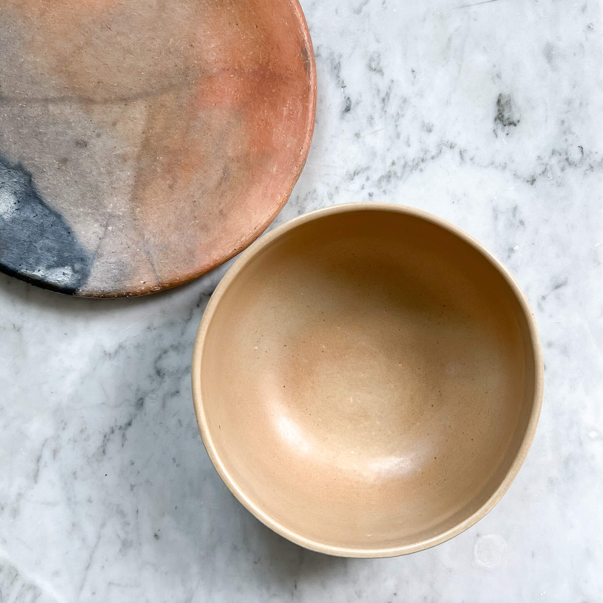 An orange clay plate from Oaxaca next to a sandy beige stoneware serving bowl on a white marble counter.