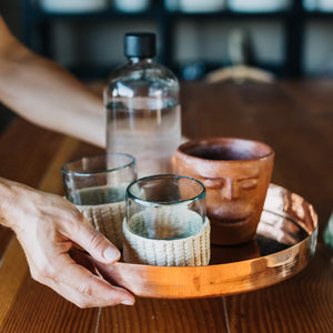 Round copper tray with palm wrapped glassware, a large clay cup and a water bottle with black cap.
