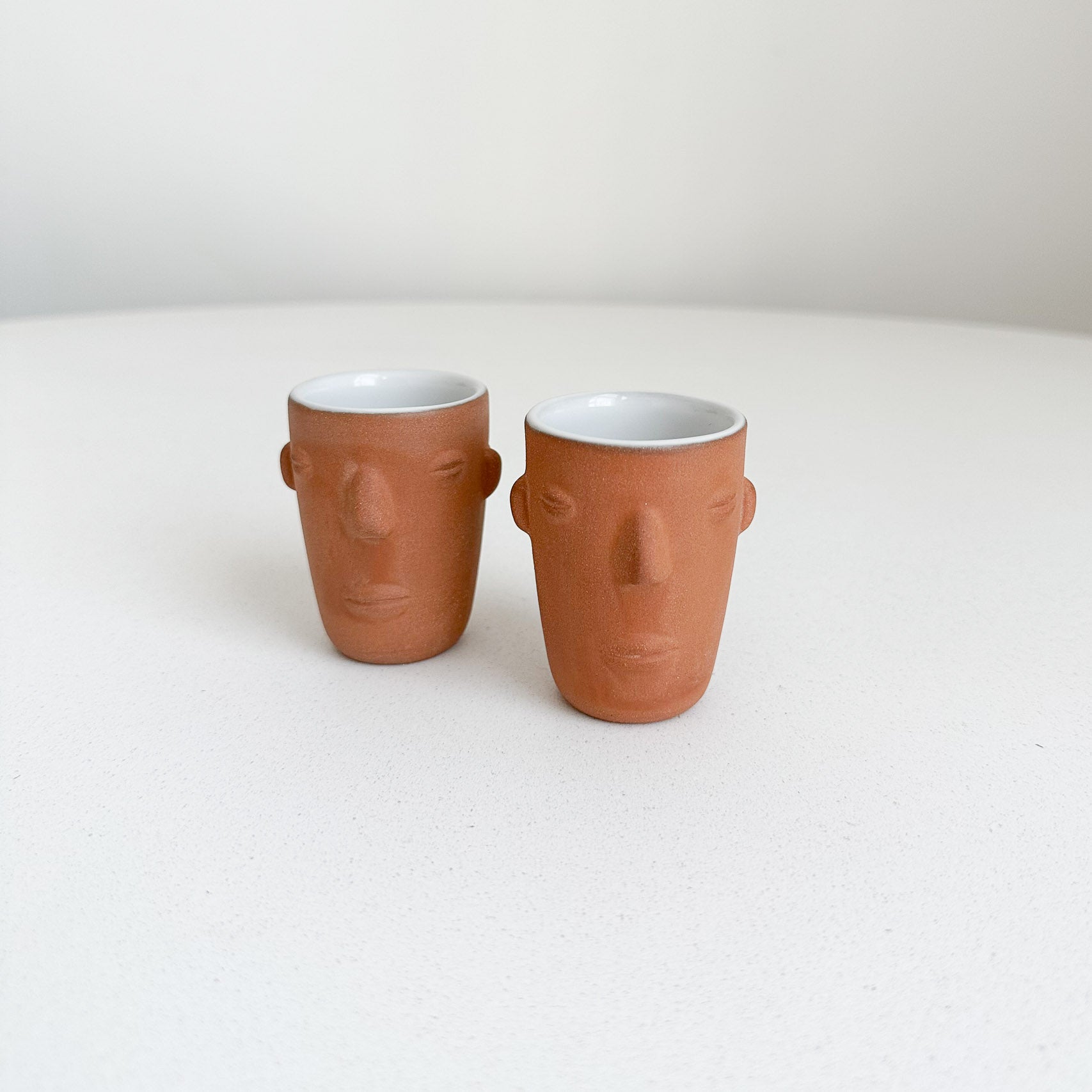 A set of two terracotta copitas with faces on a white table.
