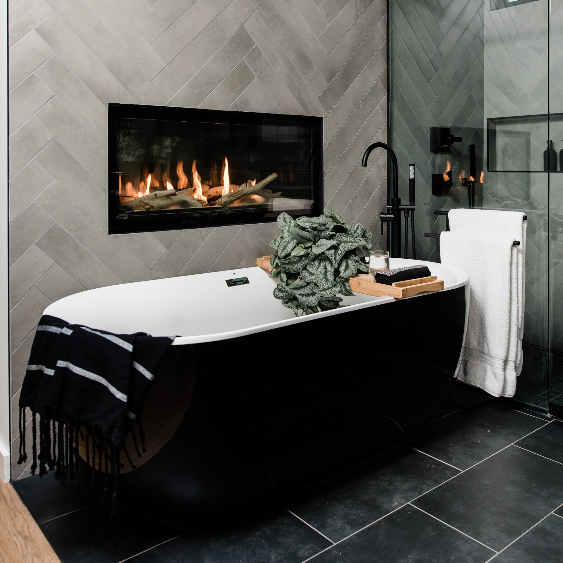 A large soaking tub draped with a black cotton towel, and a bath tray with a towel, plant and candle.