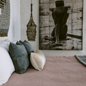 A pair of grey suede pillows along with a mini leather lumbar pillow are arranged on a bed. A hanging pendant light and large photograph are in the background.