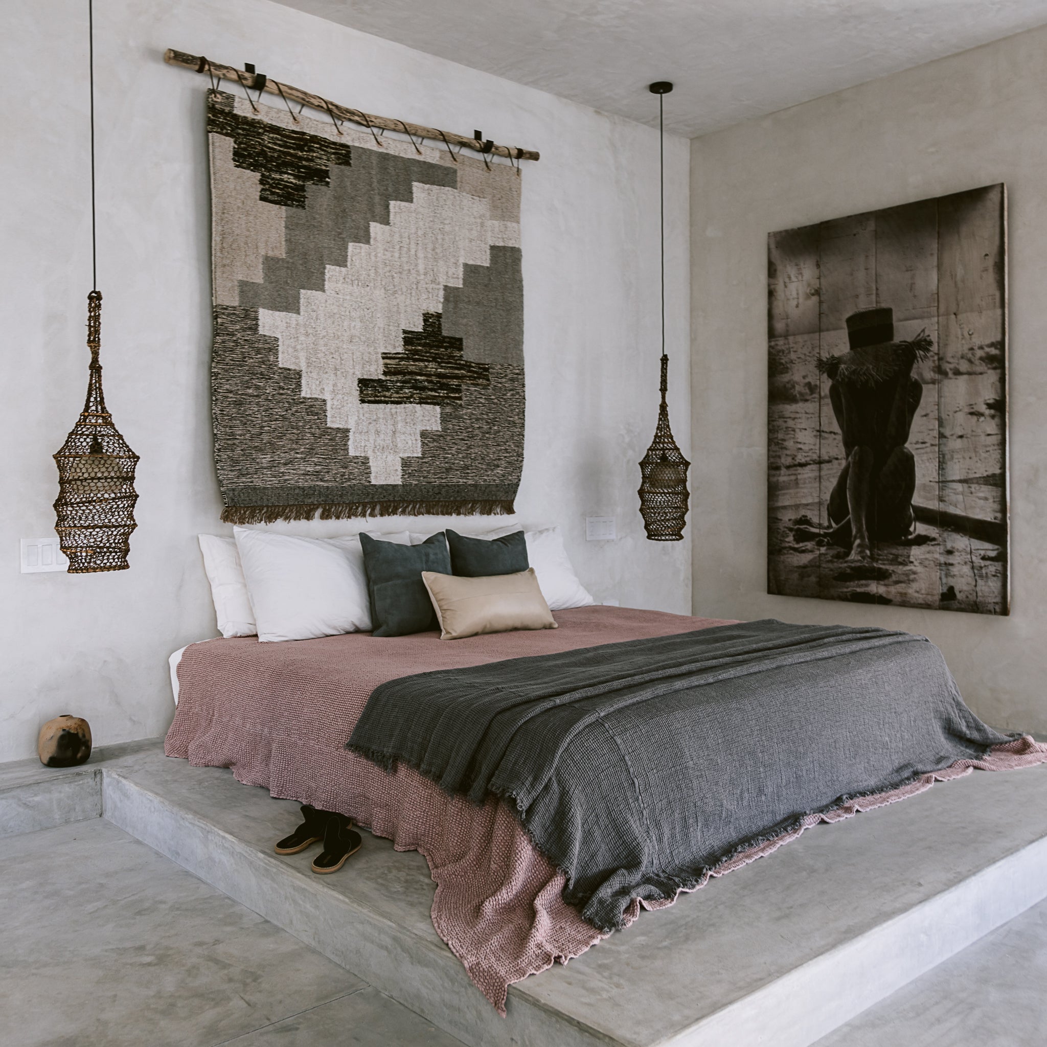 A bedroom scene in a modern beachhouse includes a concrete platform bed, plush bedding, leather throw pillows, a decorative wall-hanging rug and a large portrait of a woman. 