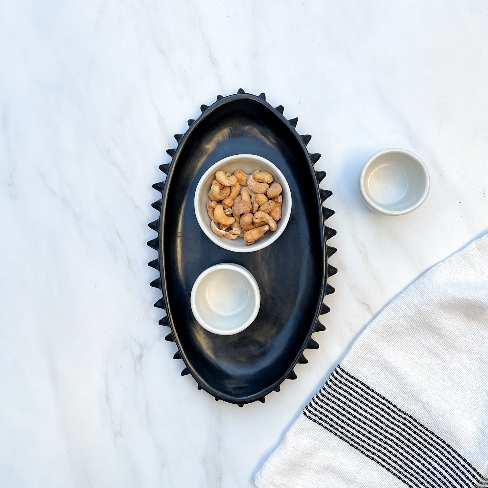 A Oaxaca black clay tray with a spiky design holding a mezcal copita, and a bowl of nuts. A cream hand towel is alongside.