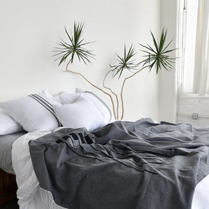A bed made with a gray handwoven cotton throw blanket, cotton euro sham pillows and a white throw pillow. A tall yucca plant stands alongside.