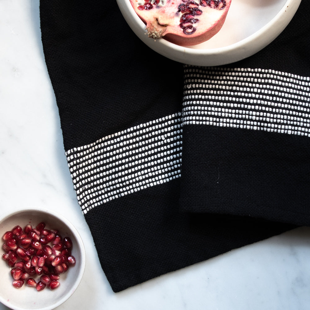Black cotton hand towel with white stripes on a white marble counter with pomegranates in white bowls.