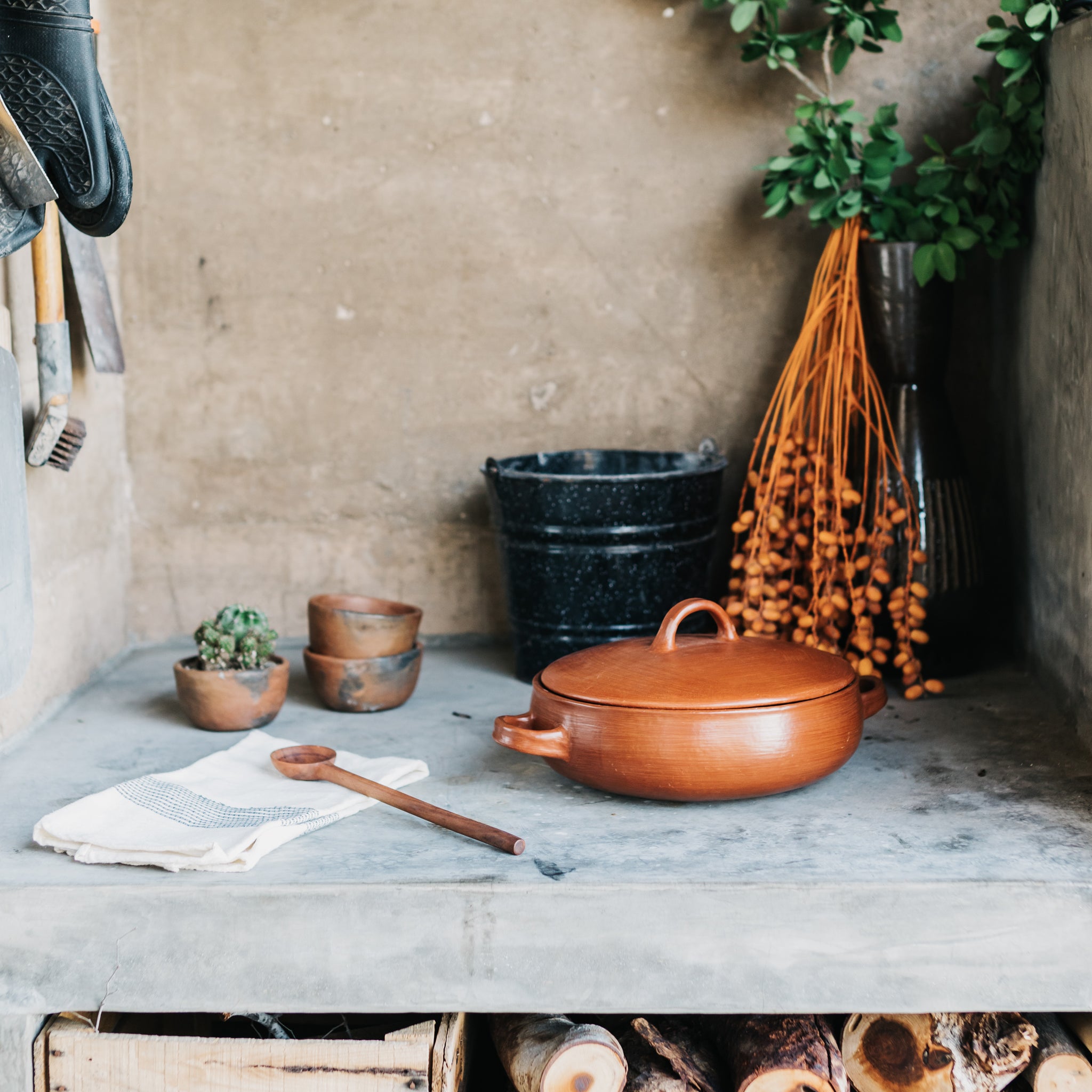 An outdoor wood-fired oven with a Oaxaca red clay cazuela, a set of stacked red clay bowls, a serving spoon and cotton hand towel.