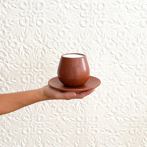 An outstretched hand holding a Oaxaca red clay mug with plate.