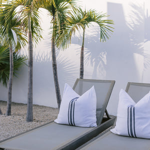 Lounge chairs with white extra-large throw pillows are next to palm trees.