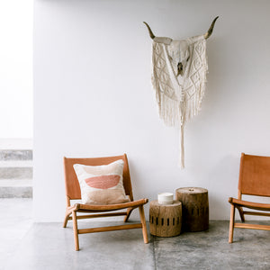 A corner in a modern beach house including two matching leather chairs, a beige and cream wool throw pillow, two side tables and a wall-hanging cattle skull adorned with macrame.