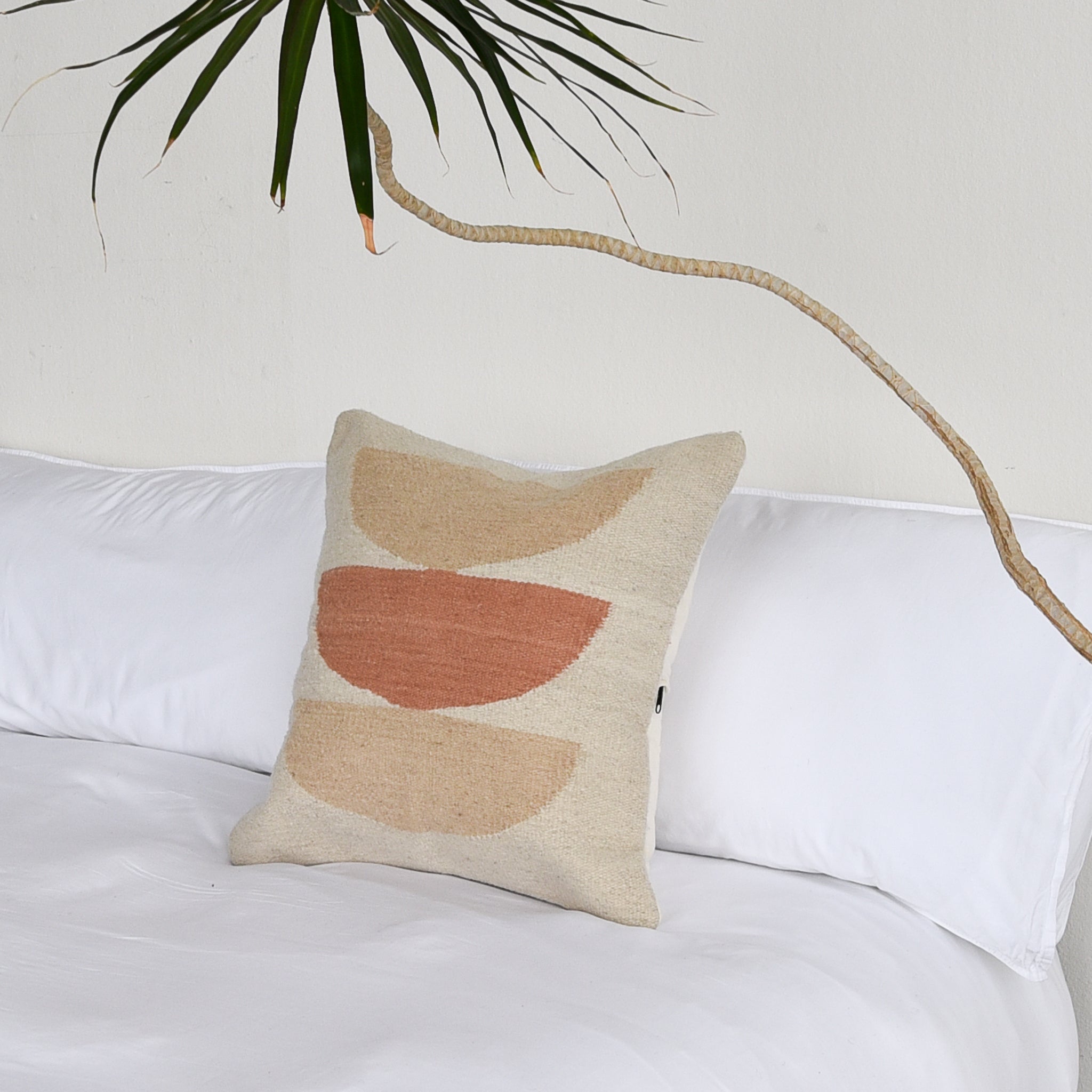 A wool throw pillow in desert colors on a white bed with the branch of a yucca overhanging the pillow.