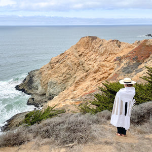 A woman standing on the edge of the California coast wrapped in the San Borja Mexican blanket and wearing a palm sombrero hat.