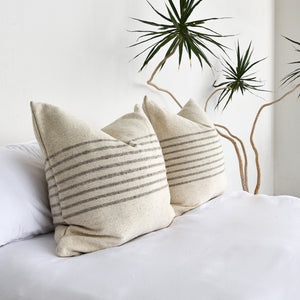 A pair of wool large throw pillows on a bed with all white bedding. A large yucca indoor plant is next to the bed.
