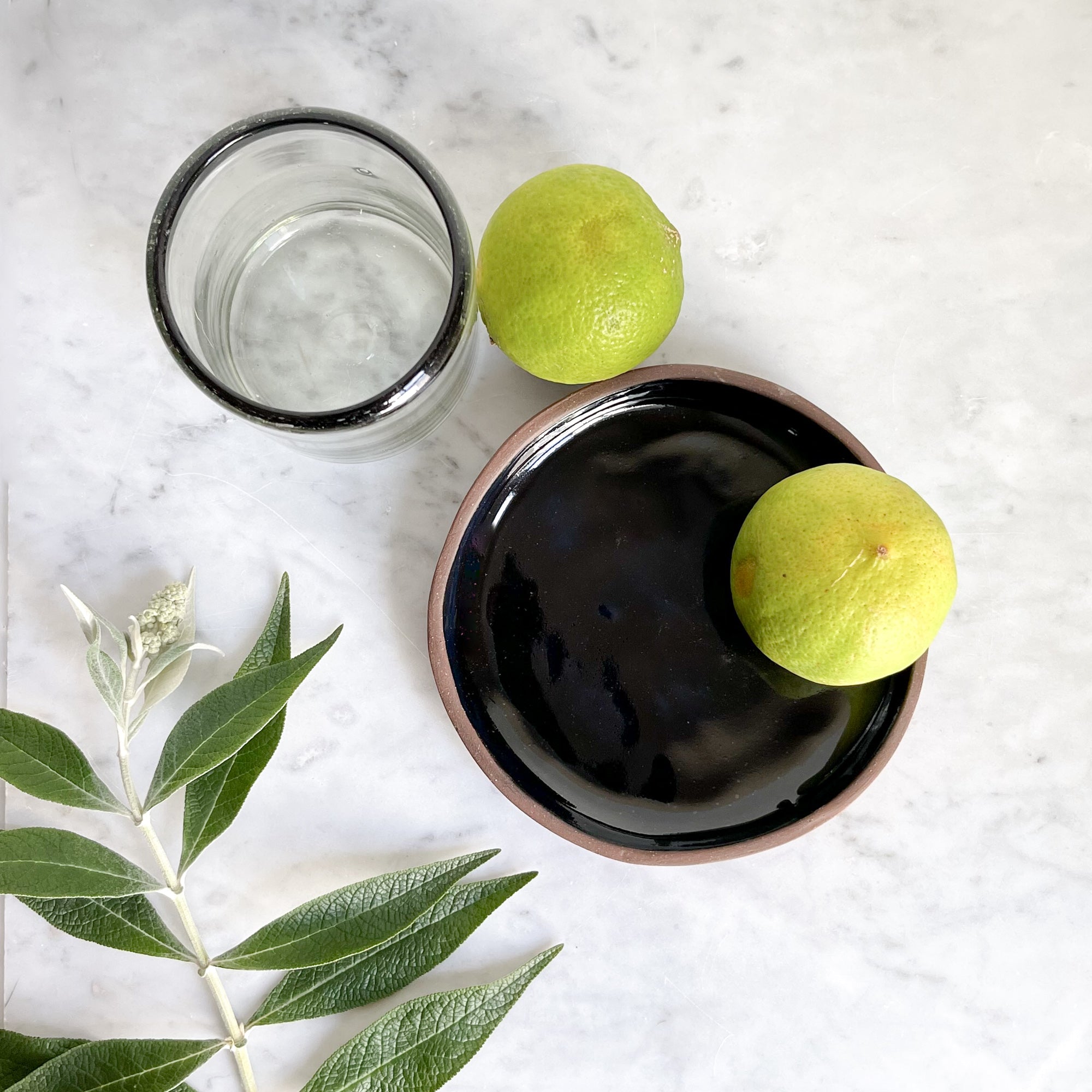 A black terra-cotta plate with limes, next to a small black-rimmed glass.
