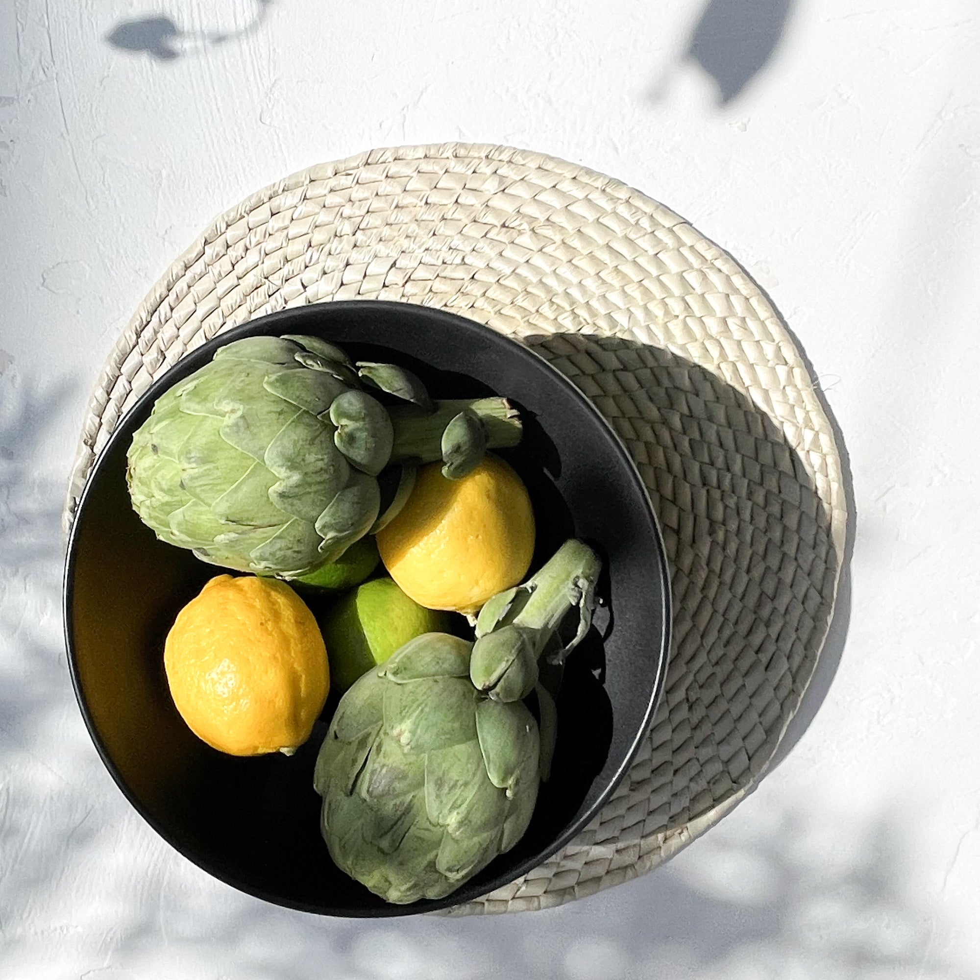 Matte black stoneware ceramic serving bowl with artichokes, fruit and a handwoven palm placemat.