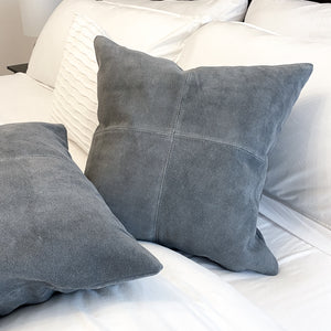 A pair of grey suede throw pillows on a bed with white sheets and blankets.