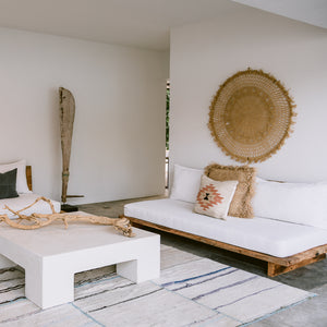 The living room of a modern beach house features white, minimalist furniture, palm accents, driftwood and handwoven wool pillows and a rug.