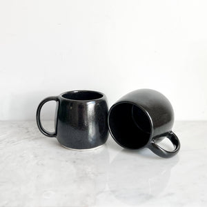 Two black stoneware ceramic mugs, one laid on it's side.