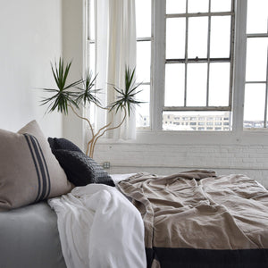 An urban loft scene featuring a bed, bright windows, and Oaxaca textile bedding along with a black braided leather throw pillow.